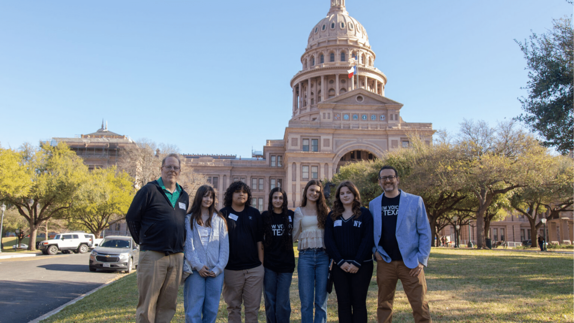 Seven people from New Voices Texas — two advisors and five high schoolers — stand in front of the Texas Capitol building.