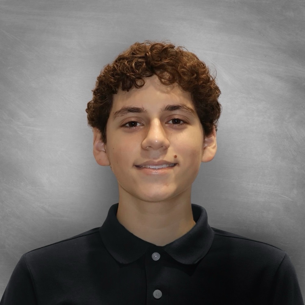 A pale-skinned teenage boy with brown curly hair wearing a black polo smiles at the camera.
