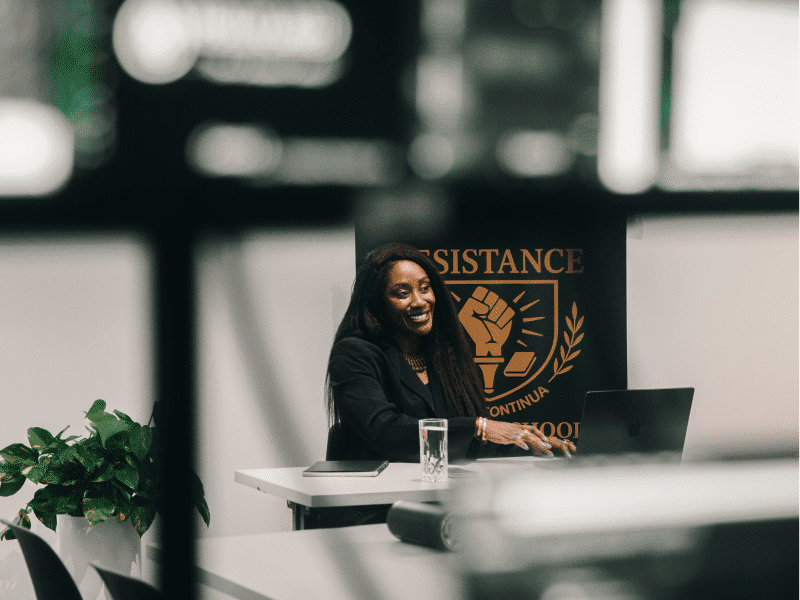 Karen Attiah, a Black woman with relaxed hair swept over her shoulder, is smiling and looking away from the camera. She is wearing a black blazer and top and sitting in front of a laptop. Behind her sits a banner for Attiah's Resistance Studies Series, with an upraised fist that says Resistance.
