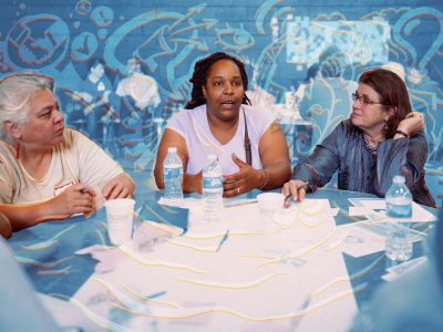 Three women of different races sit at a table discussing. There is a light blue overlay with orange-outlined transparent icons like lightbulbs, a speech bubble with a microphone, a handshake and more.