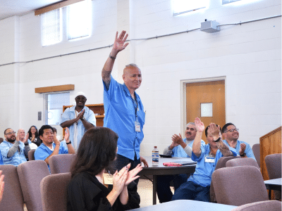 A Latino man with a mohawk in a blue uniform smiles and waves amid a crowd of multiracial audience members in San Quentin's blue uniforms and business-casual dress.
