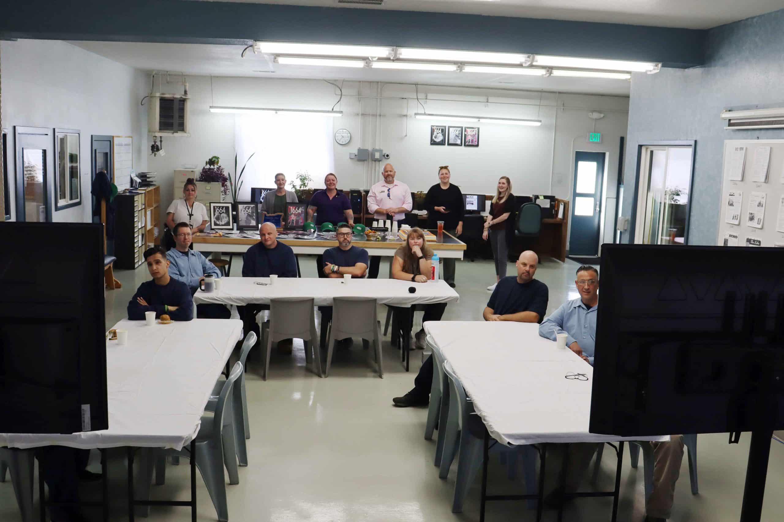 Thirteen members of 1664 magazine and The Echo face two monitors that project the 2025 American Penal Press Awards ceremony. A table with a cake decorated with a 1664 magazine cover and issues of the publications is at the back.