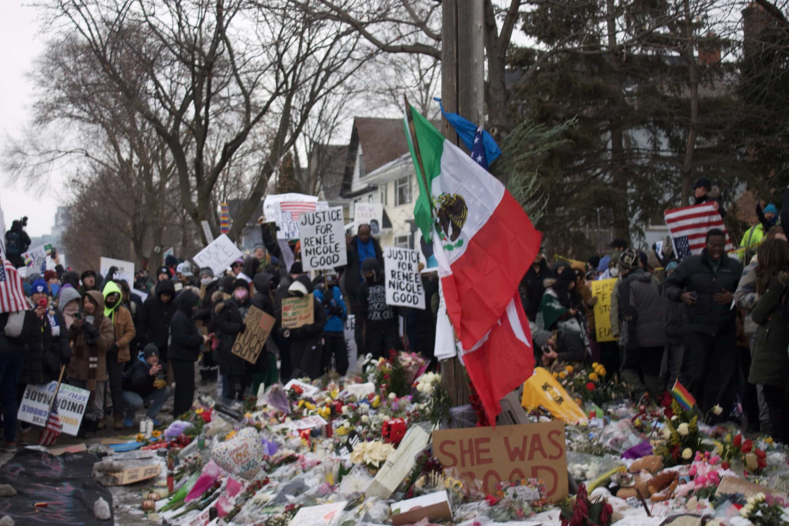A crowd of onlookers gathers around a memorial covered in flower bouquets. At the center are a Mexican flag and American flag, grounded by a sign that says "She Was Good." Some onlookers hold signs that read "Justice for Renee Nicole Brown."