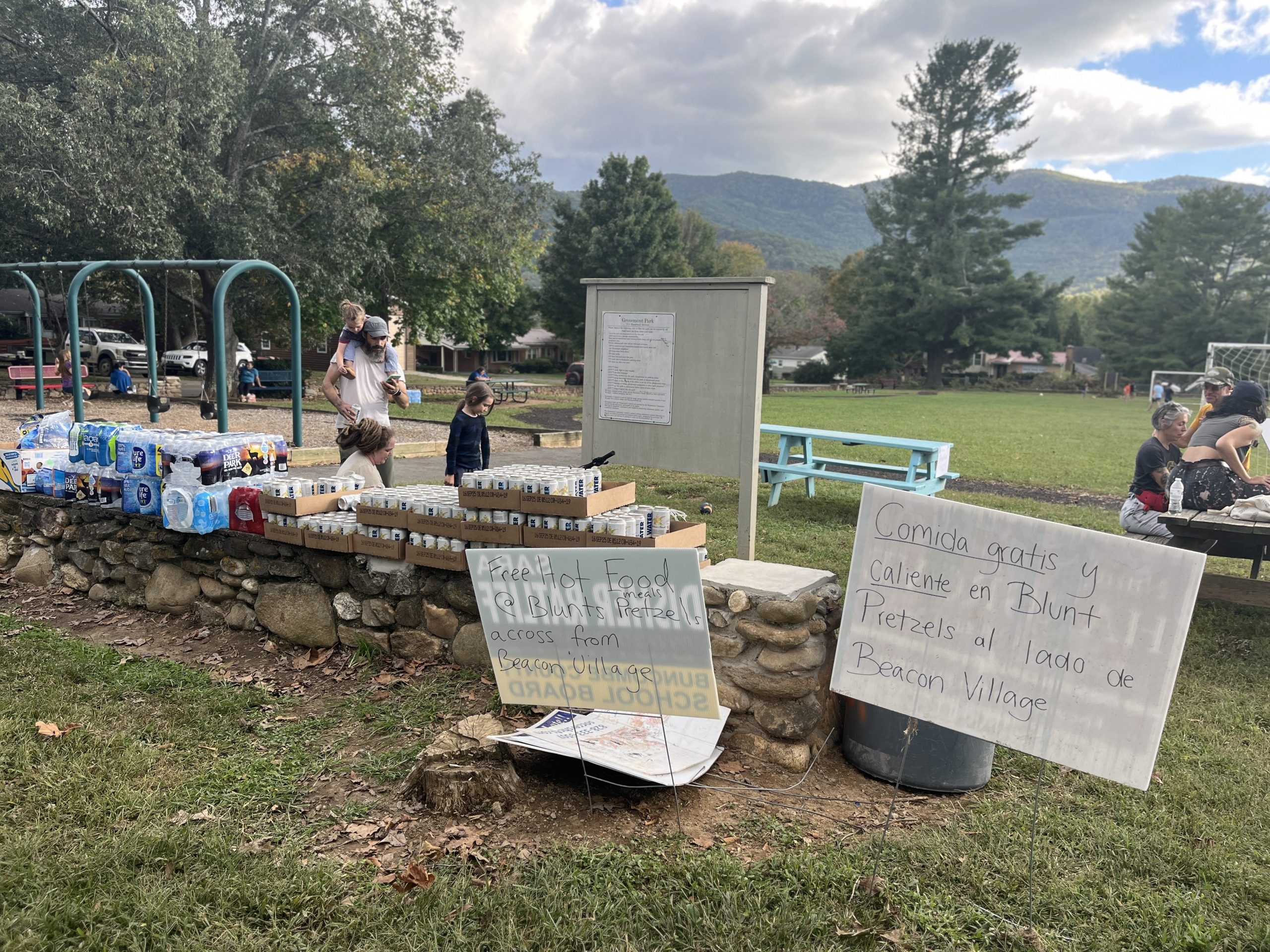 Families congregate around free drinks at Government Park after Hurricane Helene; two signs in English and Spanish advertise free hot food and meals at Blunts Pretzels across from Beacon Village.