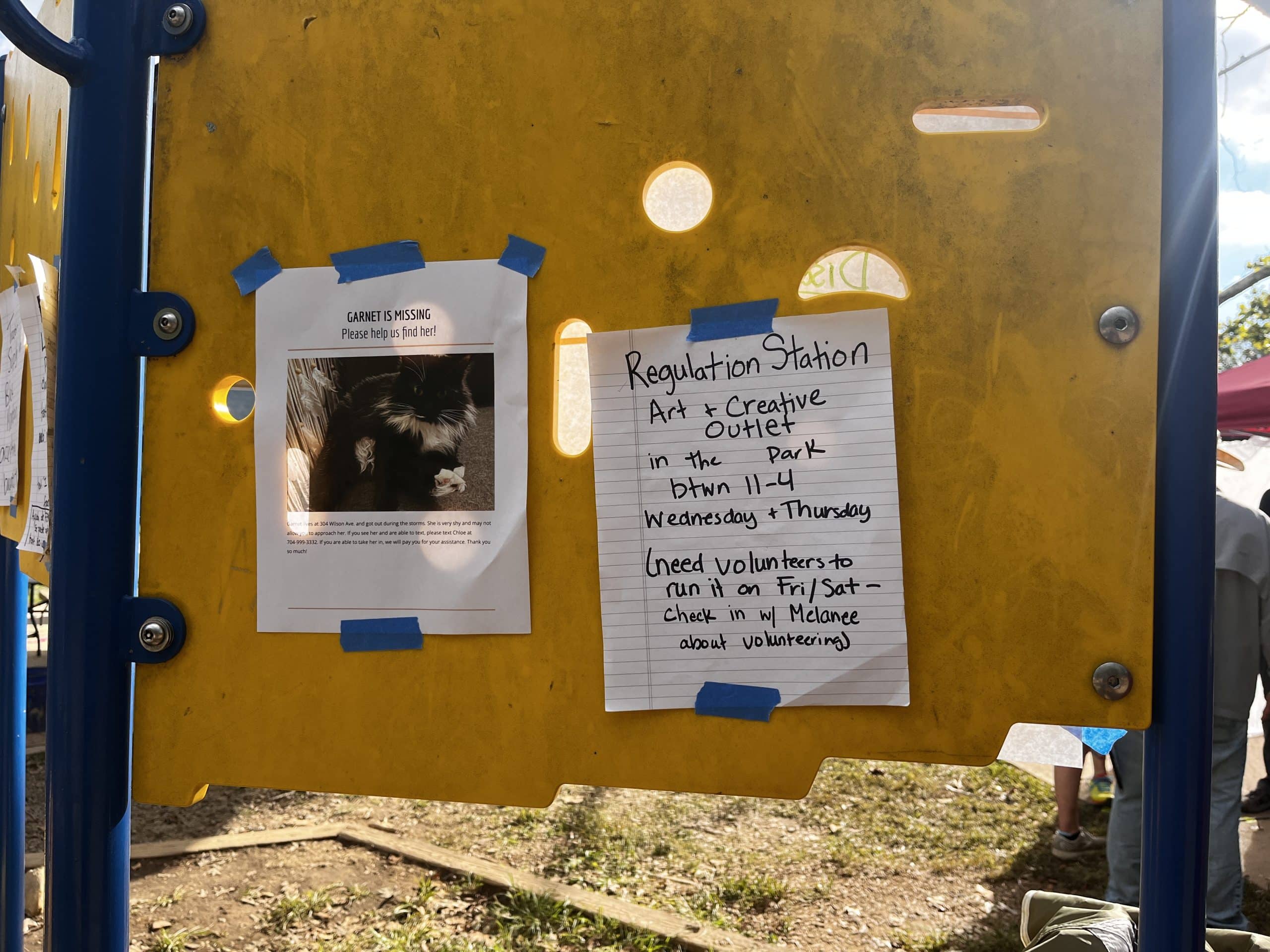 A back of a sign being used as a bulletin board, with two signs taped up: One advertising Garnet, a missing cat, and another on a sheet of lined paper that says: Regulation Station, Art + Creative Outlet in the park between 11-4 Wednesday + Thursday (need volunteers to run it on Fri/Sat - check in w/ Melanee about volunteering)