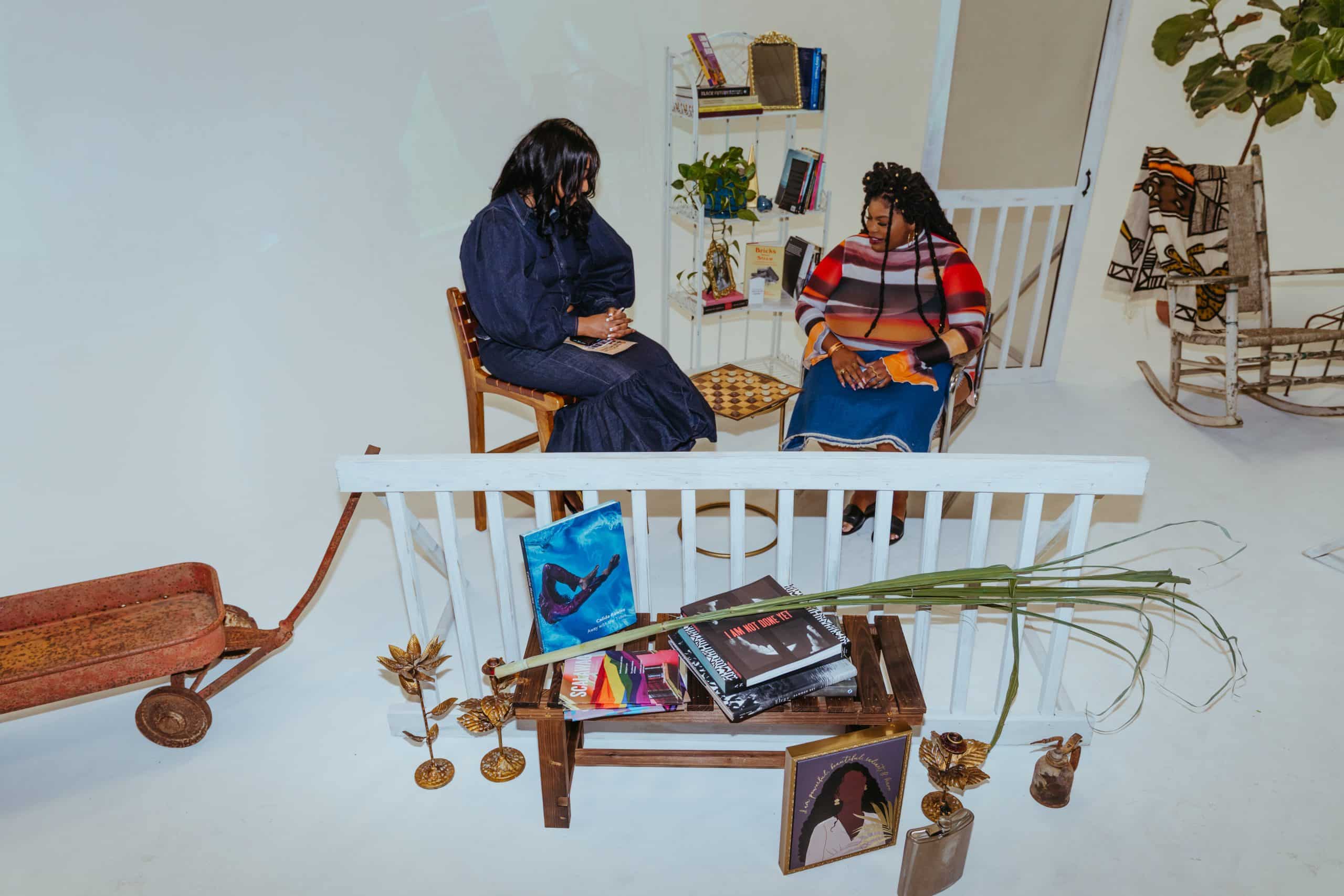 Diamond Hardiman, a Black woman with relaxed, curled hair, is in a denim two-piece set with a balloon-sleeve top and trumpet skirt next to Venneikia Williams, a Black fem whose hair is in half-up, half-down twists. Williams wears a striped top and denim skirt. The two are situated around a checkerboard, in the middle of a display of books by Black authors.