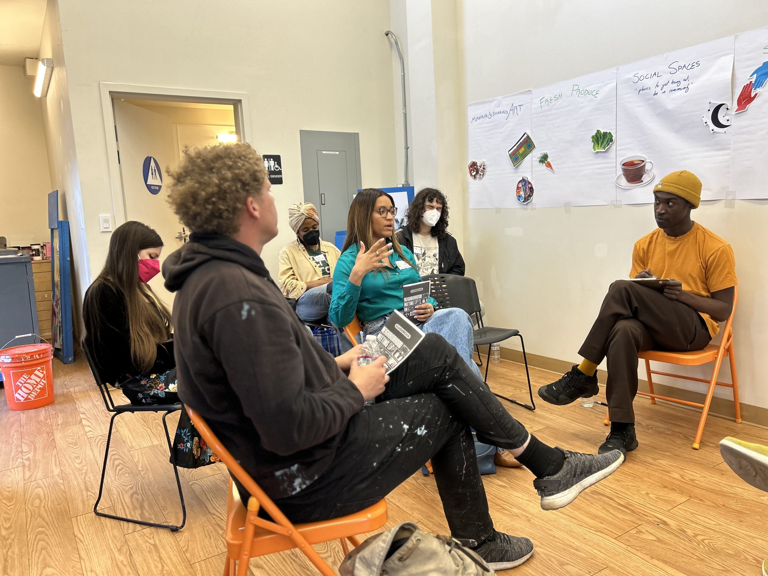 Six people of different races sit in a circle talking in front of a set of brainstorming posters about art, fresh produce, and social spaces.