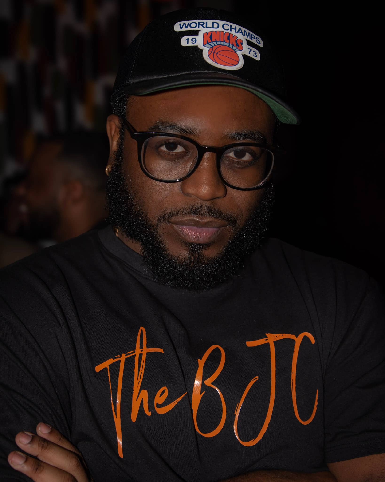 Headshot of JaJuan Malachi, a Black man with glasses and a beard who wears a serious expression and a shirt that says The BJC, along with a World Champs Knicks 1973 cap.
