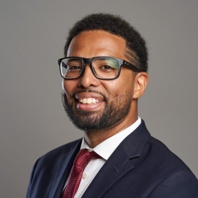 Headshot of Michael Butler, who has rectangular glasses and is wearing a suit and red tie. He is smiling at the camera and has slightly-grown out natural hair.
