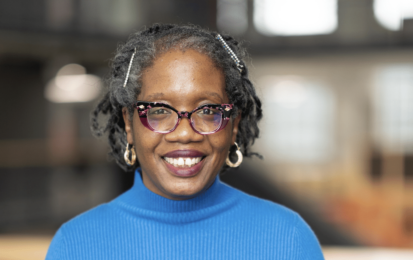 Adrienne Johnson Martin's headshot. She is a middle-aged Black woman with braids pinned back, a blue turtleneck, pink cat-eye glasses, and gold hoop earrings smiling at the camera.