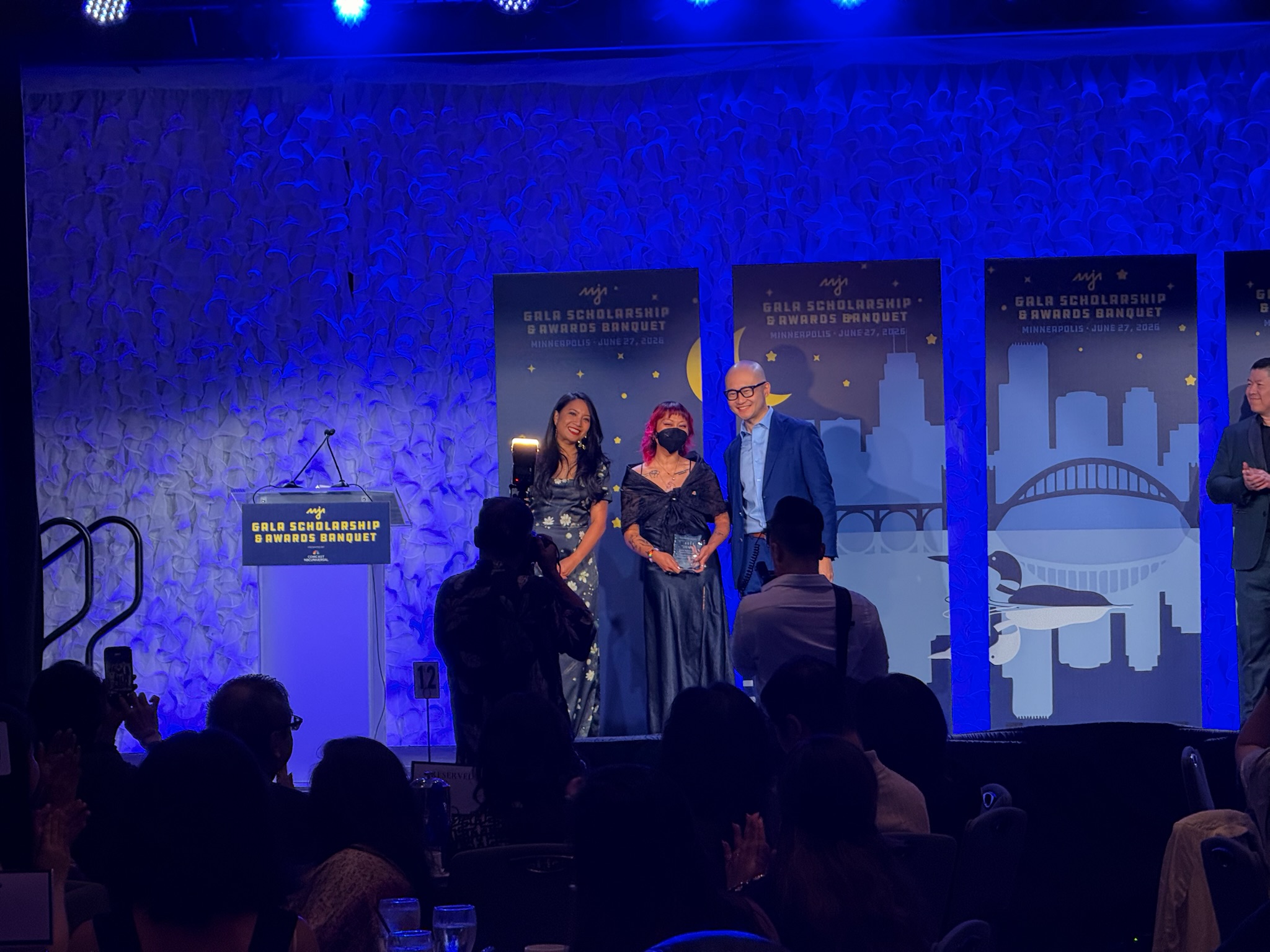 Co-director James Salanga, a Filipino-Chinese person with pink curled hair, stands in a black alampay, watermelon pin, and black floor-length dress while holding up the 2026 AAJA Community Award for Leadership in Diversity and Solidarity. They are flanked by outgoing AAJA board members Nicole Dungca and Frank Bi, and all three are on stage at the AAJA 2026 Gala Scholarship & Awards Banquet.