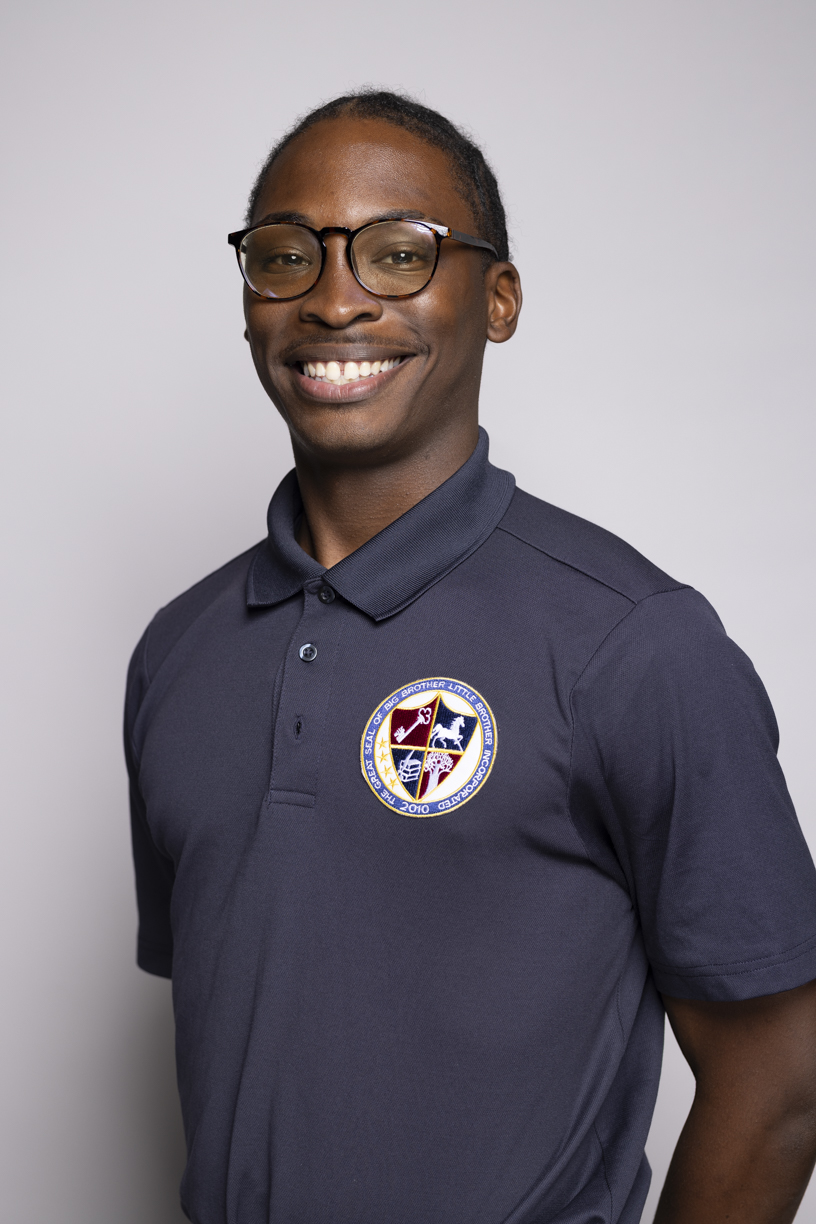 Headshot of Jonathan Charles, a young Black man with close-shaven hair who smiles at the camera. He wears glasses and a gray-blue polo with a Big Brother Little Brother patch.