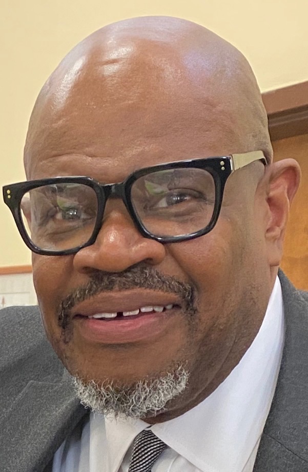 Photo of James Ford, a middle-aged Black man with bald hair, square glasses, and a short silver goatee. Ford is wearing a suit and looks, mid-smile, at the camera.