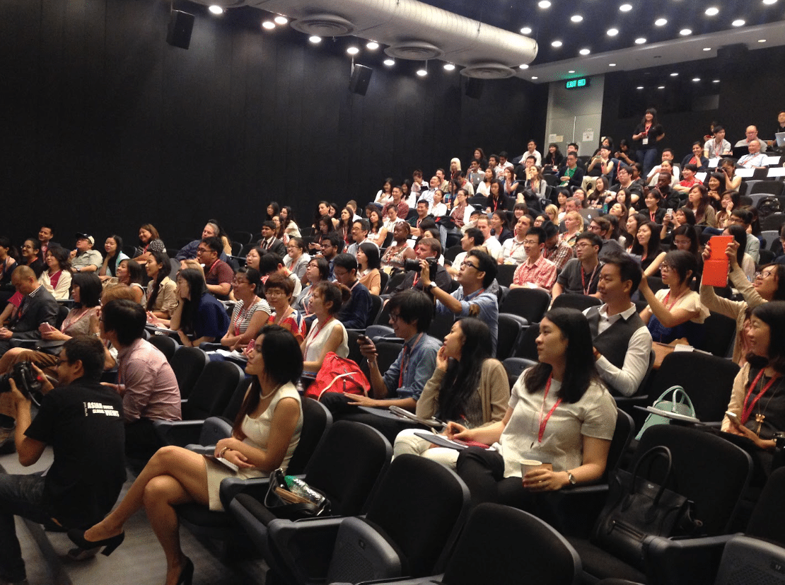 A full crowd of dozens of predominantly Asian journalists across ethnicities and genders fills a theater/auditorium-like room. Several are taking photos at the front.
