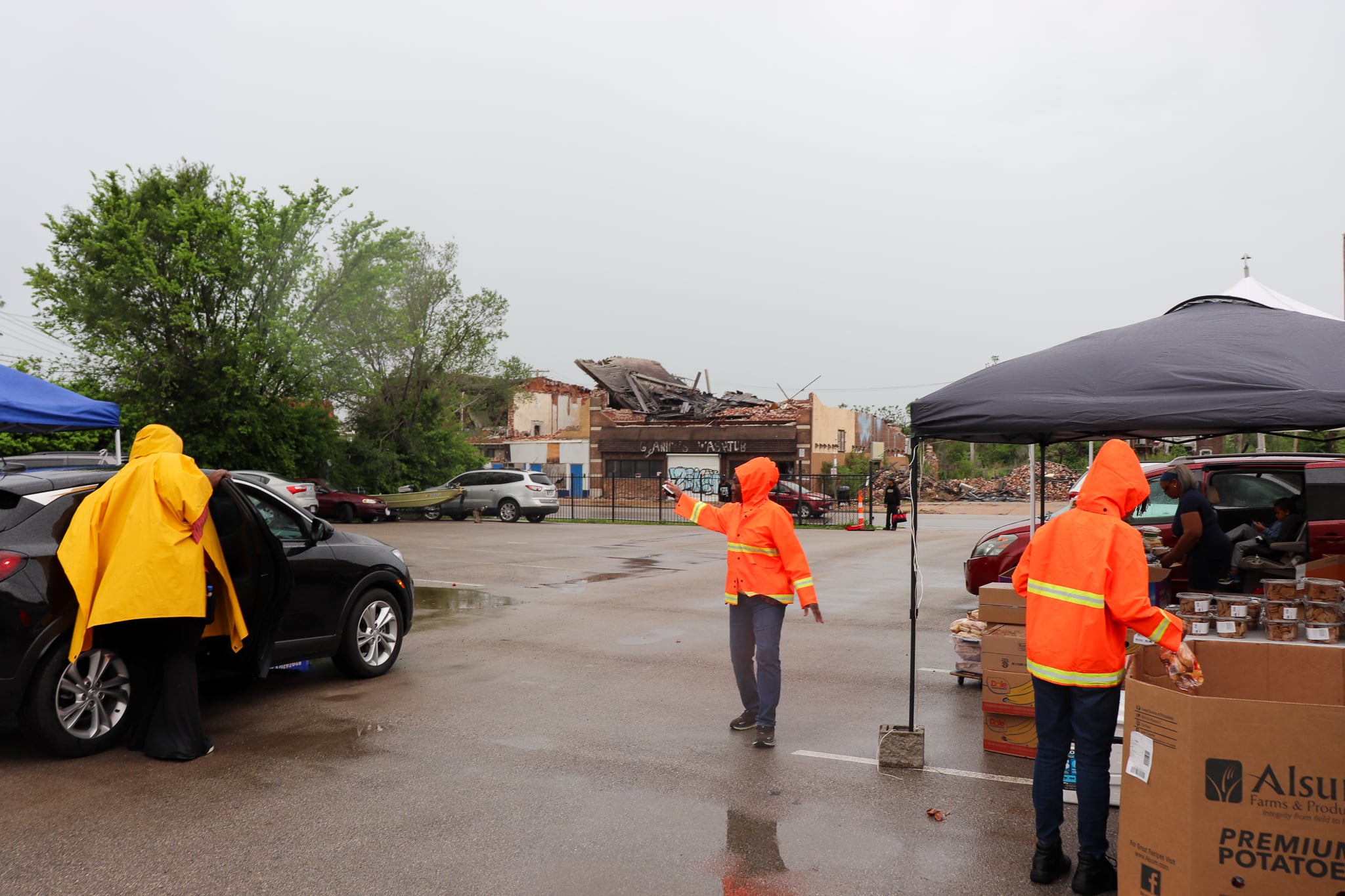 Volunteers in orange raincoats direct distribution of food in front of tornado-stricken buildings on a rainy day. 